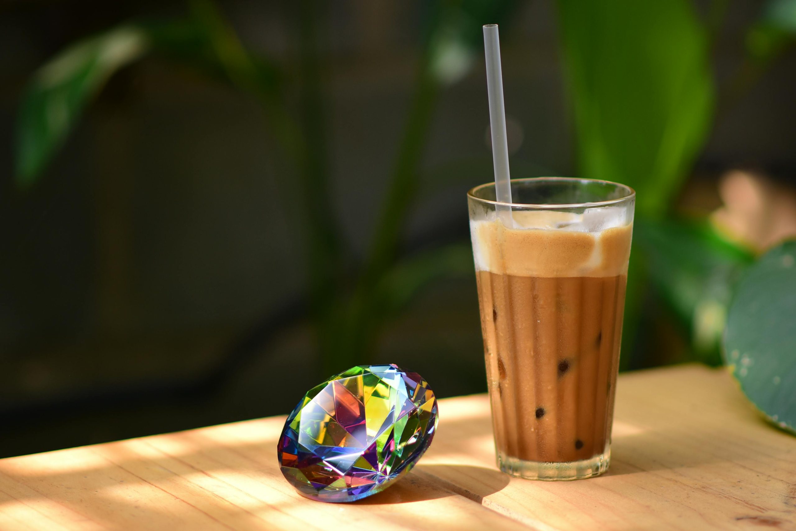 Glass of iced coffee and colorful gem on wooden table in sunlight, ideal for lifestyle and beverage themes.